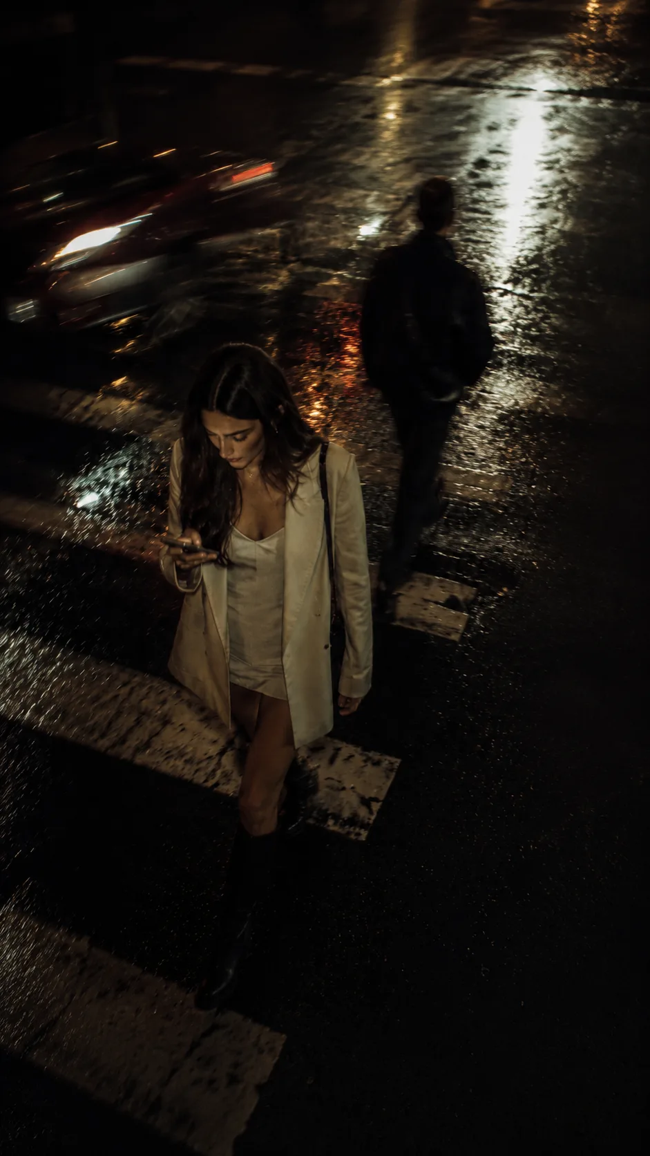 A woman alone on a wet city street at night, lit by her phone.