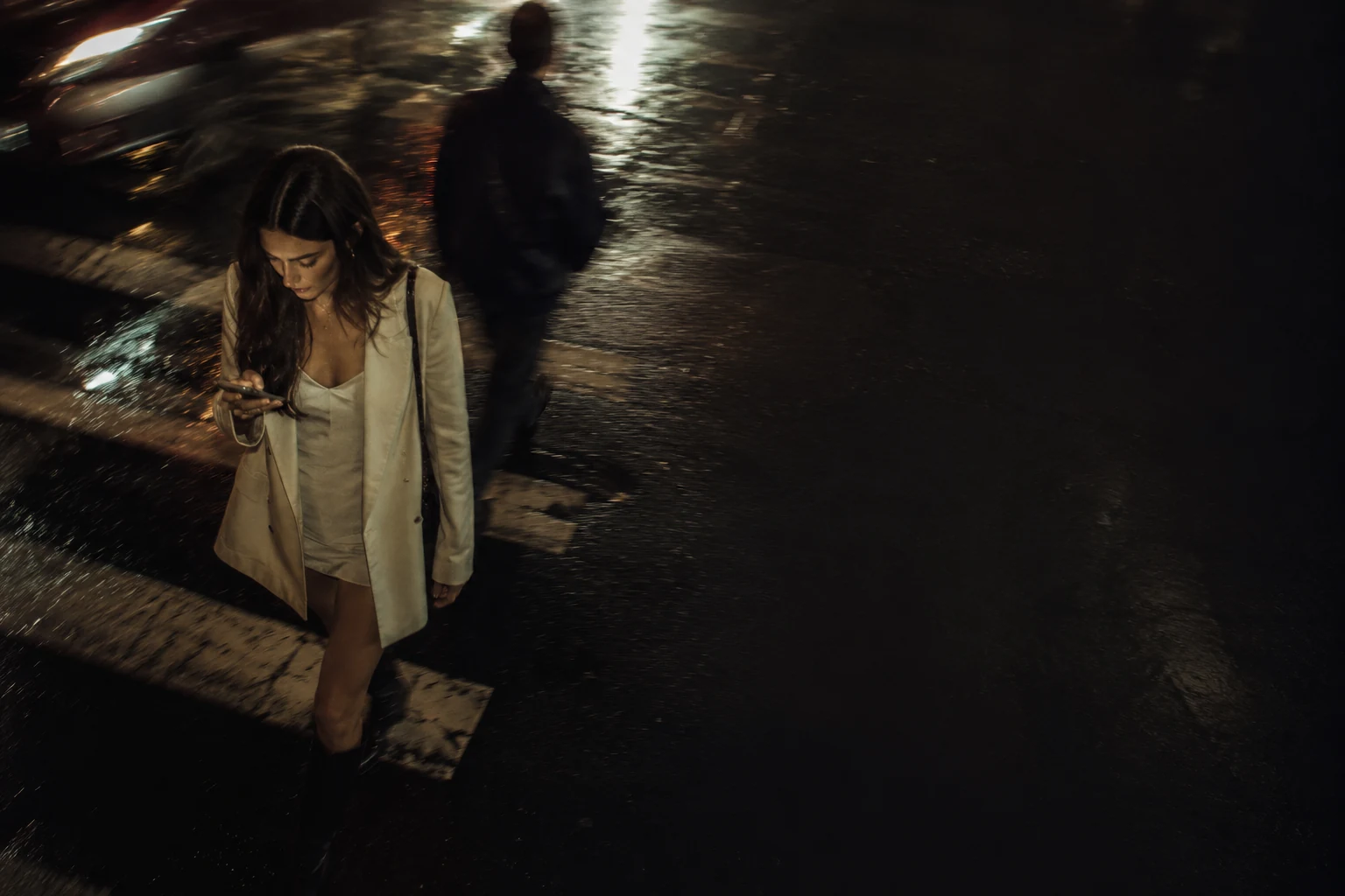 A woman alone on a wet city street at night, lit by her phone.